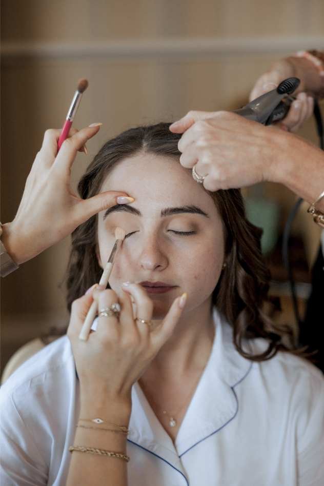 A woman with closed eyes sits calmly as two people apply makeup to her eyelids and style her wavy brown hair with a curling iron. She wears a light blue top, preparing for a session with a top newborn photographer.