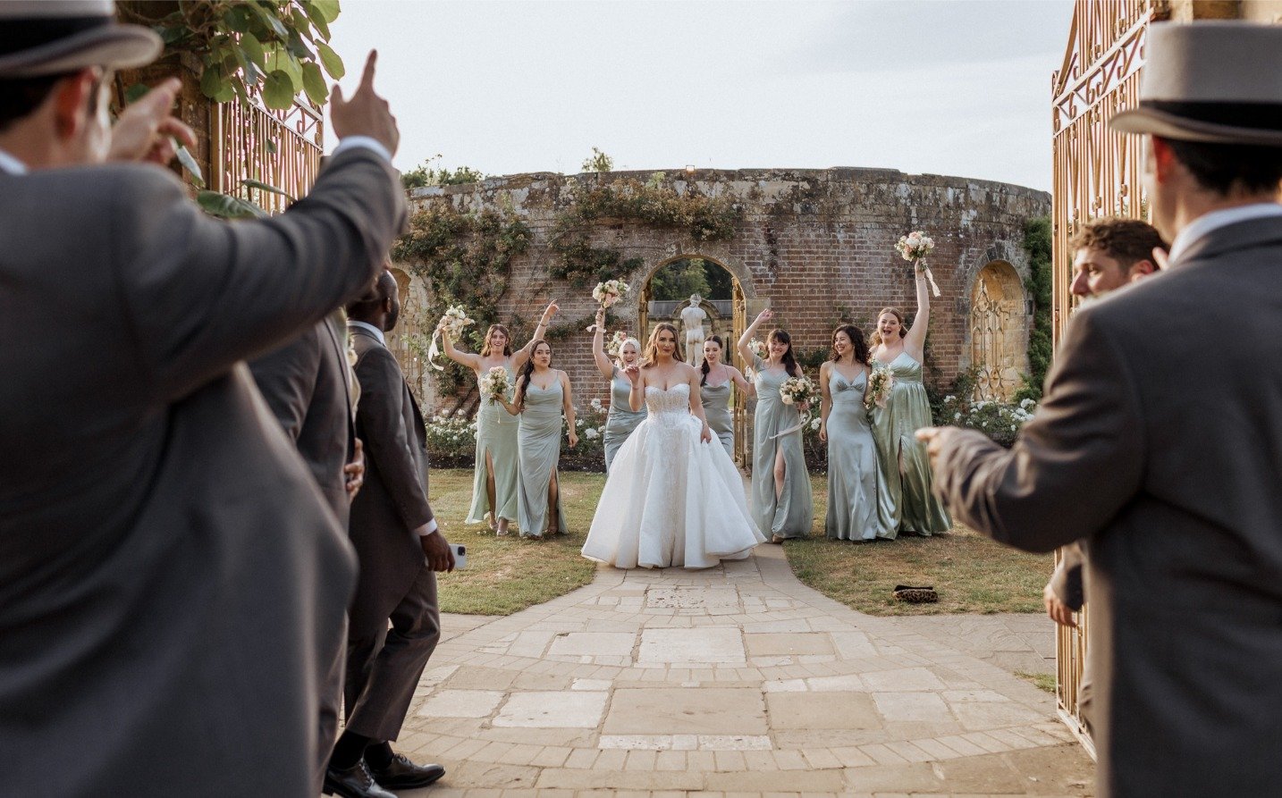A bride in a white dress walks down a stone path at a Hever Castle wedding, surrounded by bridesmaids in green holding bouquets. Groomsmen in gray suits and hats cheer from both sides, with a rustic brick wall as the backdrop.
