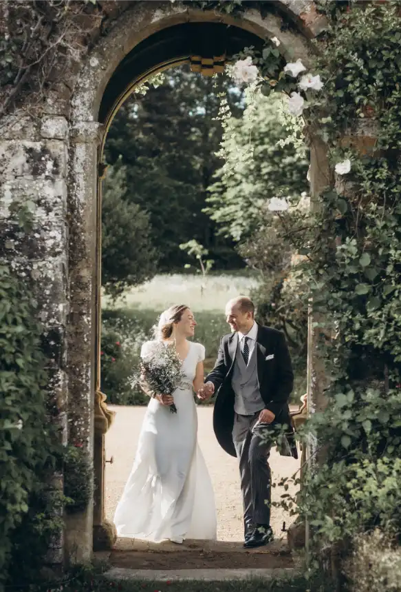 Home 2 A bride and groom smile at each other while walking hand in hand through a flower-covered stone archway in a lush garden on their wedding day, perfectly captured by a Kent wedding photographer at this luxury wedding venue in Kent.