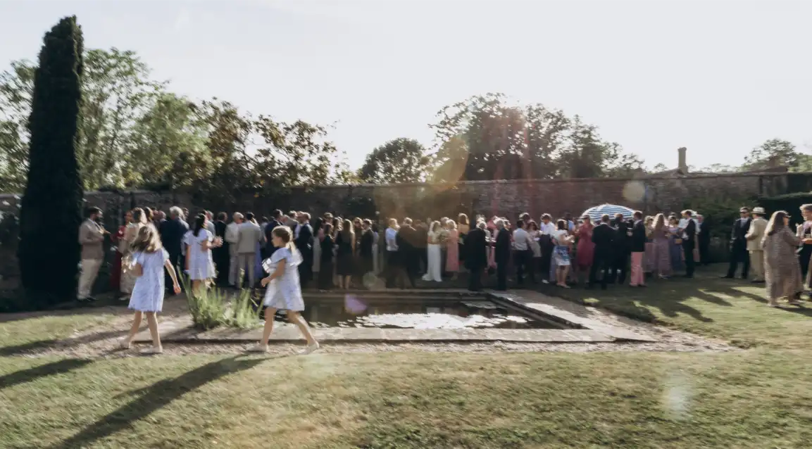 A large group of people stands and mingles near a rectangular pond in a sunny garden, while two women in light dresses walk across the grass in the foreground, capturing the joyful atmosphere of a wedding at Hever Castle.