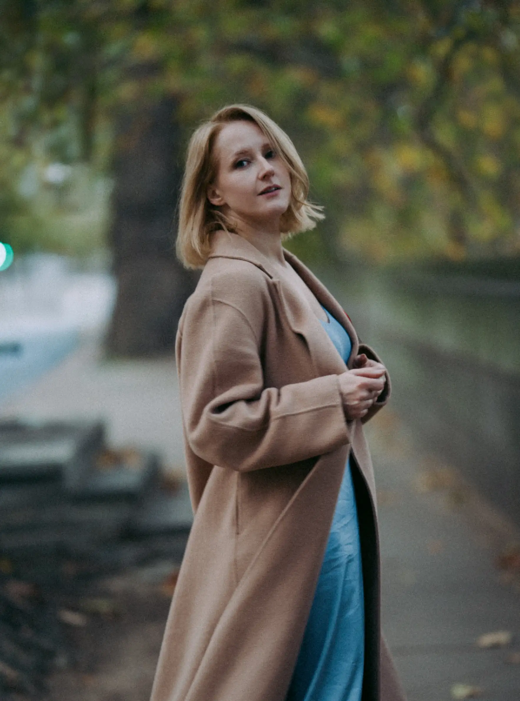 Home 3 A woman in a tan coat and blue dress stands on a sidewalk, looking back over her shoulder. Captured in an elegant, editorial style by a Surrey Wedding Photographer, the background is beautifully blurred with green trees and a stone wall.